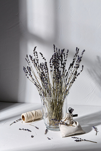 aromatherapy and organic concept - still life of dried lavender flowers in glass vase, craft sachet bag and rope dropping shadows on white surface