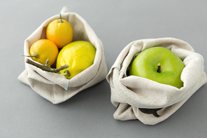 consumerism and eco friendly concept - fruits in reusable canvas bags for food shopping s on grey background