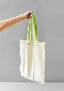 consumerism and eco friendly concept - hand holding reusable canvas bag for food shopping on grey background