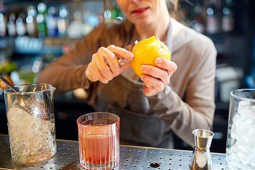 alcohol drinks, people and luxury concept - woman bartender with glass and peeler removing peel from orange and preparing cocktail at bar
