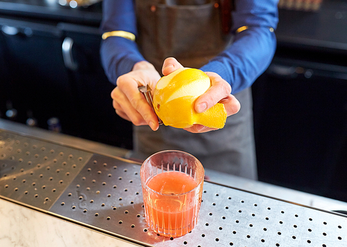 alcohol drinks, people and luxury concept - bartender with glass and peeler removing peel from lemon and preparing cocktail at bar