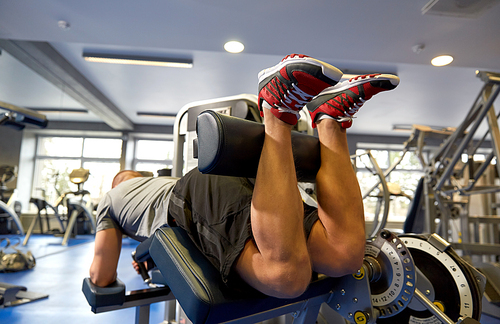 sport, fitness and bodybuilding concept - man exercising and flexing muscles on leg curl machine in gym