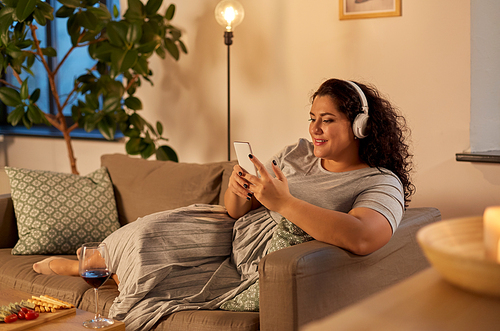 technology, people and leisure concept - happy young woman in headphones listening to music on smartphone at home in evening