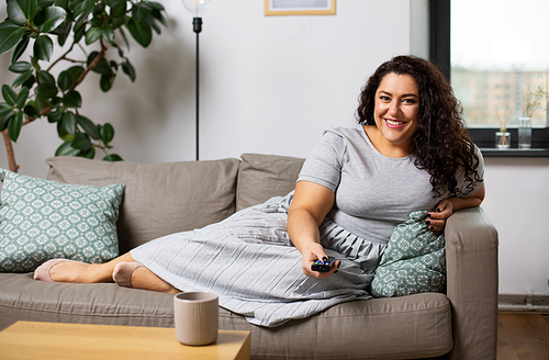 people and leisure concept - happy young woman lying on sofa with remote control and watching tv at home