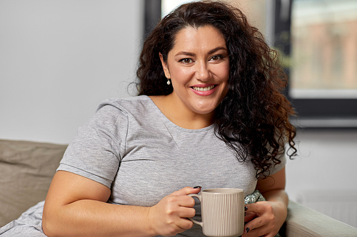 people and leisure concept - happy young woman drinking coffee or tea on sofa at home