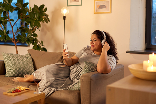 technology, people and leisure concept - happy young woman in headphones listening to music on smartphone at home in evening