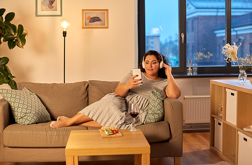 technology, people and leisure concept - happy young woman in headphones listening to music on smartphone at home in evening