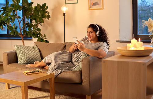 technology, people and leisure concept - happy young woman in headphones listening to music on smartphone at home in evening