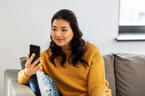 people, technology and leisure concept - happy smiling asian young woman in yellow sweater with smartphone sitting on sofa at home