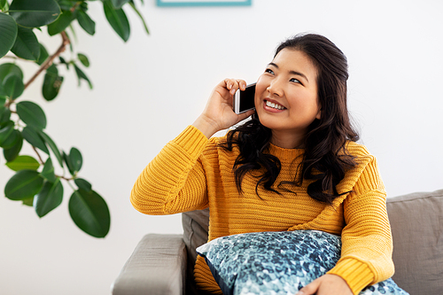 people, technology and communication concept - happy smiling asian young woman in yellow sweater sitting on sofa and calling on smartphone at home