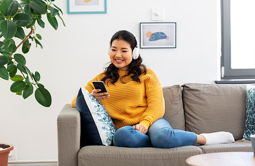 people and leisure concept - happy smiling asian young woman in wireless headphones sitting on sofa and listening to music on smartphone at home