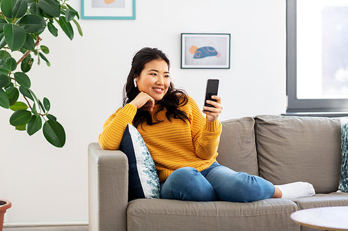 people and leisure concept - happy smiling asian young woman in wireless earphones sitting on sofa and listening to music on smartphone at home