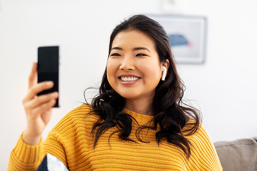 people and leisure concept - happy smiling asian young woman in wireless earphones sitting on sofa and listening to music on smartphone at home