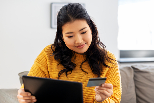 internet bank, online shopping and technology concept - happy smiling asian young woman in yellow sweater sitting on sofa with tablet pc computer and credit card at home