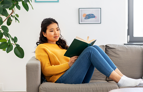 people and leisure concept - happy asian young woman in yellow sweater sitting on sofa and reading book at home