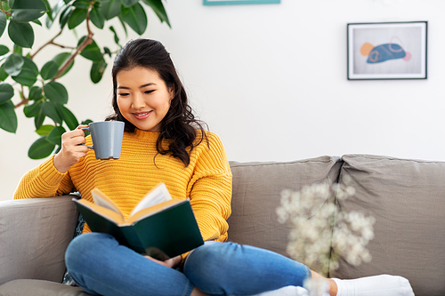 people and leisure concept - happy asian young woman in yellow sweater sitting on sofa, reading book and drinking coffee at home