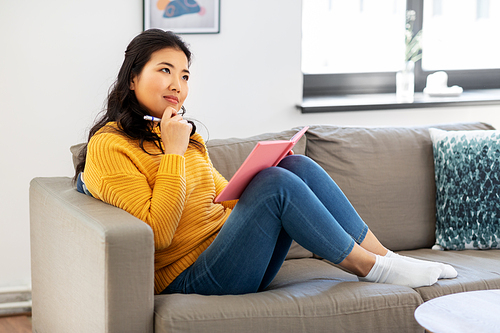 people and leisure concept - thinking asian young woman with diary or notebook sitting on sofa at home