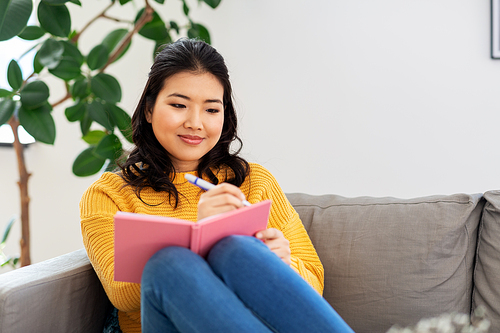 people and leisure concept - happy smiling asian young woman in yellow sweater with diary or notebook sitting on sofa at home