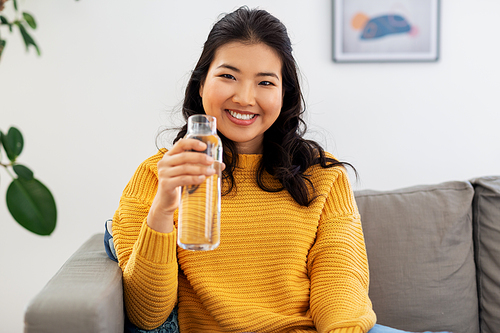 people, sustainability and leisure concept - happy smiling asian young woman in yellow sweater sitting on sofa and drinking water from glass bottle at home