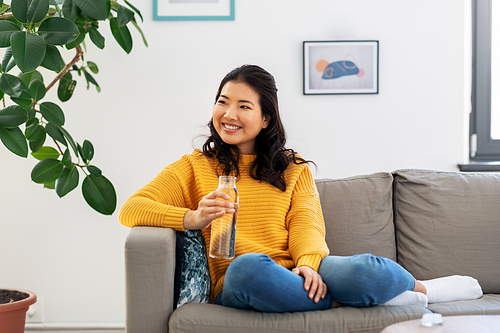 people, sustainability and leisure concept - happy smiling asian young woman in yellow sweater sitting on sofa and drinking water from glass bottle at home