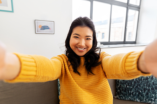 people and leisure concept - happy smiling asian young woman in yellow sweater taking selfie at home