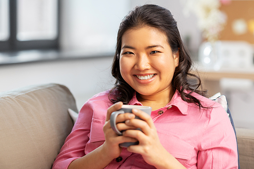 people and leisure concept - happy smiling asian young woman in pink shirt sitting on sofa and drinking coffee at home