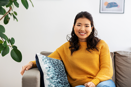 people and leisure concept - happy smiling asian young woman in yellow sweater sitting on sofa at home