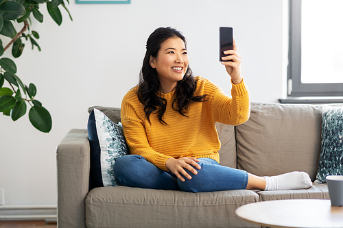 people and leisure concept - happy smiling asian young woman in yellow sweater sitting on sofa and taking selfie with smartphone at home