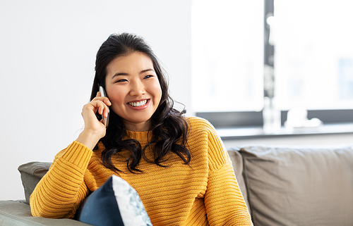 people, technology and communication concept - happy smiling asian young woman in yellow sweater sitting on sofa and calling on smartphone at home