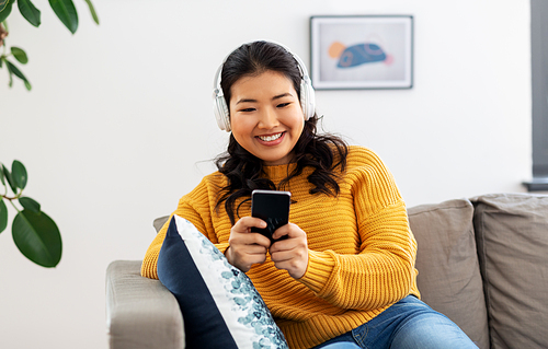 people and leisure concept - happy smiling asian young woman in wireless headphones sitting on sofa and listening to music on smartphone at home