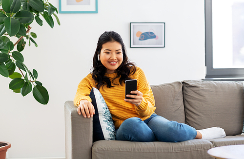 people and leisure concept - happy smiling asian young woman in wireless earphones sitting on sofa and listening to music on smartphone at home