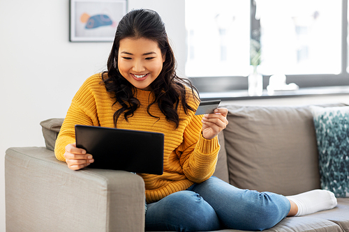 internet bank, online shopping and technology concept - happy smiling asian young woman in yellow sweater sitting on sofa with tablet pc computer and credit card at home