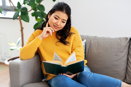 people and leisure concept - happy asian young woman in yellow sweater sitting on sofa and reading book at home