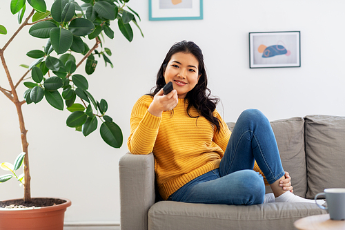 people and leisure concept - happy smiling asian young woman with tv remote control sitting on sofa at home