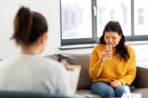 psychology and mental therapy concept - young asian woman patient drinking water and psychologist at psychotherapy session