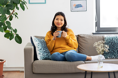 people and leisure concept - happy smiling asian young woman in yellow sweater sitting on sofa and drinking coffee at home