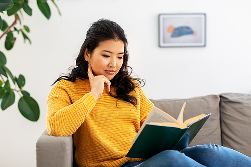 people and leisure concept - happy asian young woman in yellow sweater sitting on sofa and reading book at home