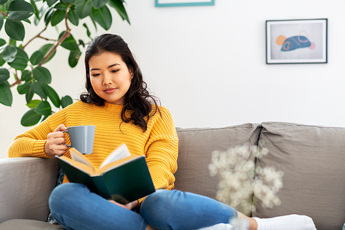 people and leisure concept - happy asian young woman in yellow sweater sitting on sofa, reading book and drinking coffee at home