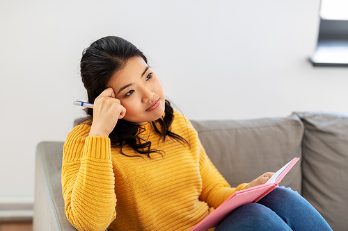 people and leisure concept - thinking asian young woman with diary or notebook sitting on sofa at home