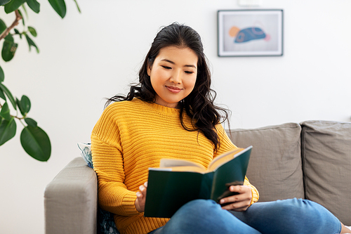 people and leisure concept - happy asian young woman in yellow sweater sitting on sofa and reading book at home