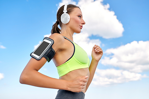 fitness, sport and healthy lifestyle concept - young woman with headphones and smartphone in armband running over blue sky