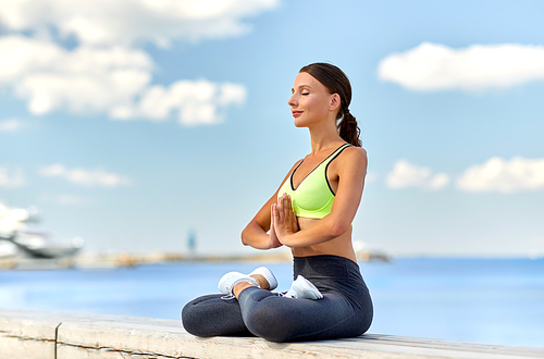 fitness, sport and yoga concept - young woman meditating in lotus pose at seaside