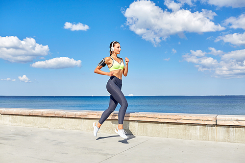 fitness, sport and healthy lifestyle concept - young woman with headphones and smartphone in armband running at seaside