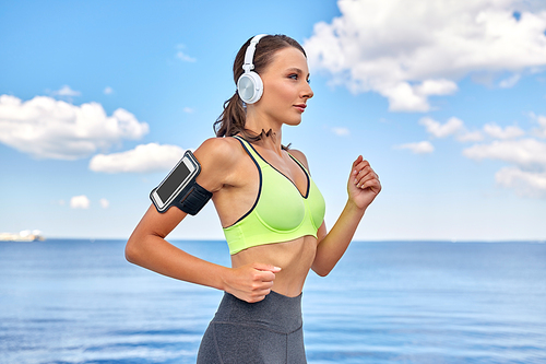 fitness, sport and healthy lifestyle concept - young woman with headphones and smartphone in armband running at seaside