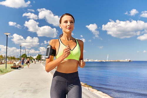 fitness, sport and healthy lifestyle concept - young woman with earphones and smartphone in arm band running along sea promenade