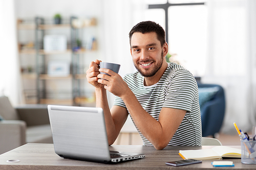 technology, remote job and lifestyle concept - happy man with laptop computer drinking coffee or tea at home office