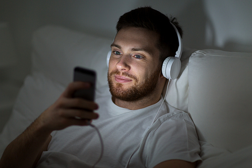 technology, internet, communication and people concept - happy smiling young man with smartphone and headphones hones listening to music in bed at night