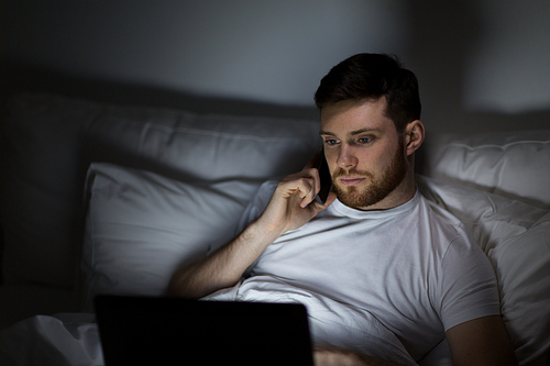 technology, internet, communication and people concept - young man in glasses with laptop computer calling on smartphone in bed at home bedroom at night