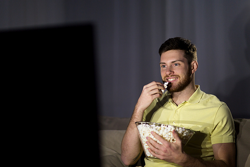 people, mass media, television and entertainment concept - happy smiling young man watching tv and eating popcorn at night at home