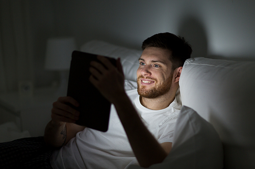 technology, internet, communication and people concept - happy young man with tablet pc computer in bed at home bedroom at night